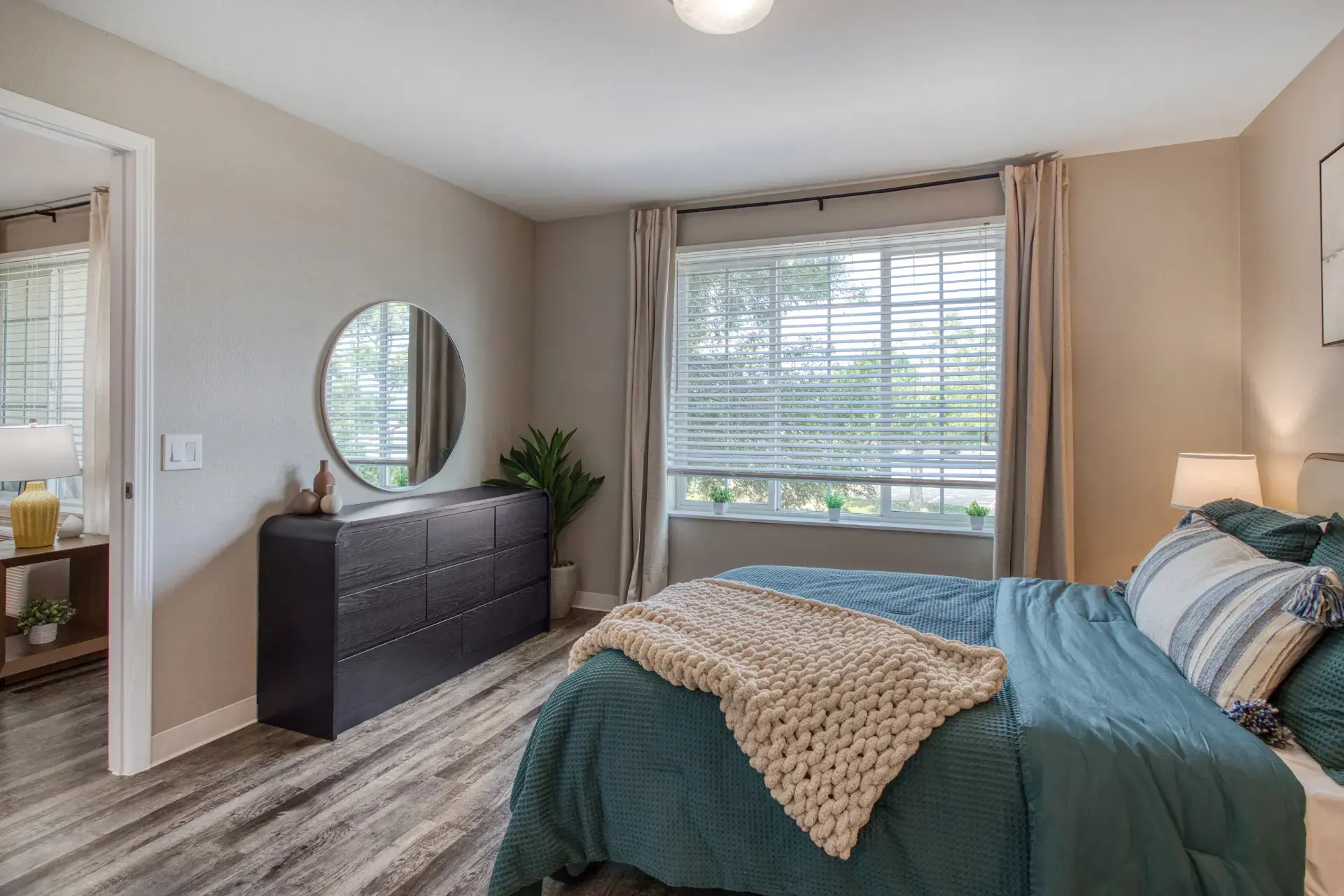 Fully furnished bedroom with circular mirror and window.